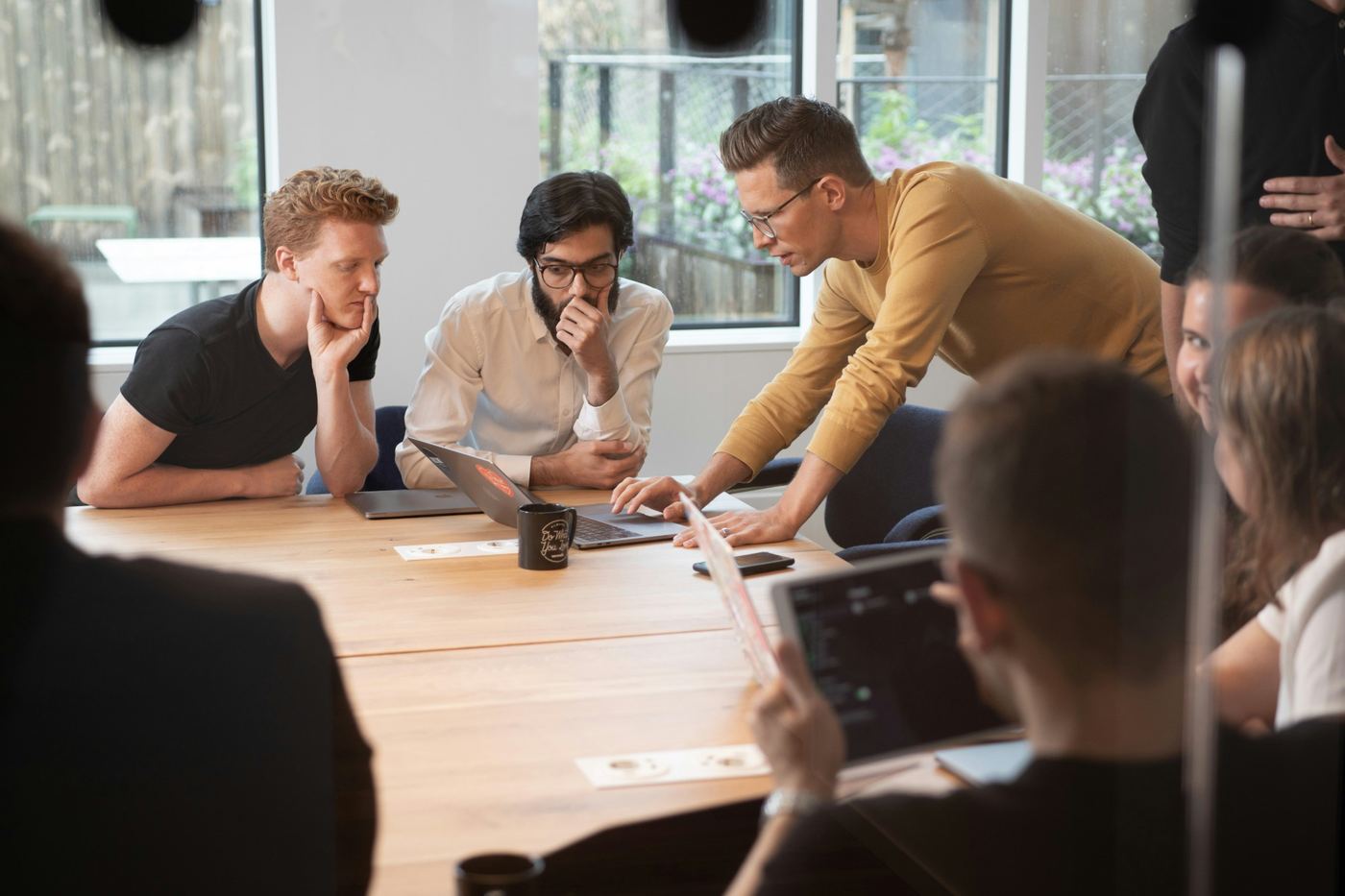 A group of people at a wooden table looking at a laptop while one of them points at the screen